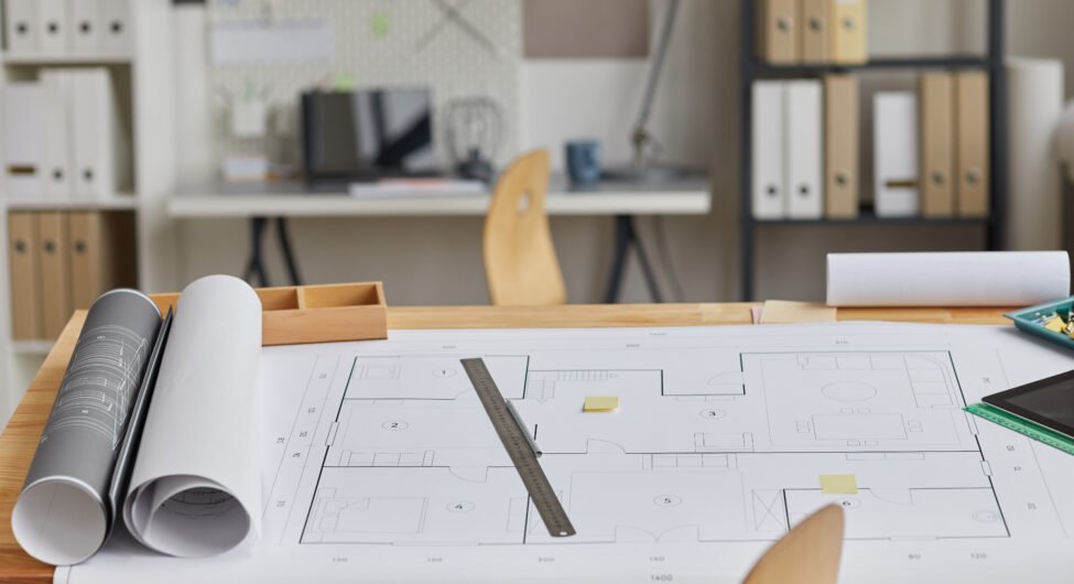 Background image of drawing table with blueprints and tools laid out in forground and architects workplace, copy space